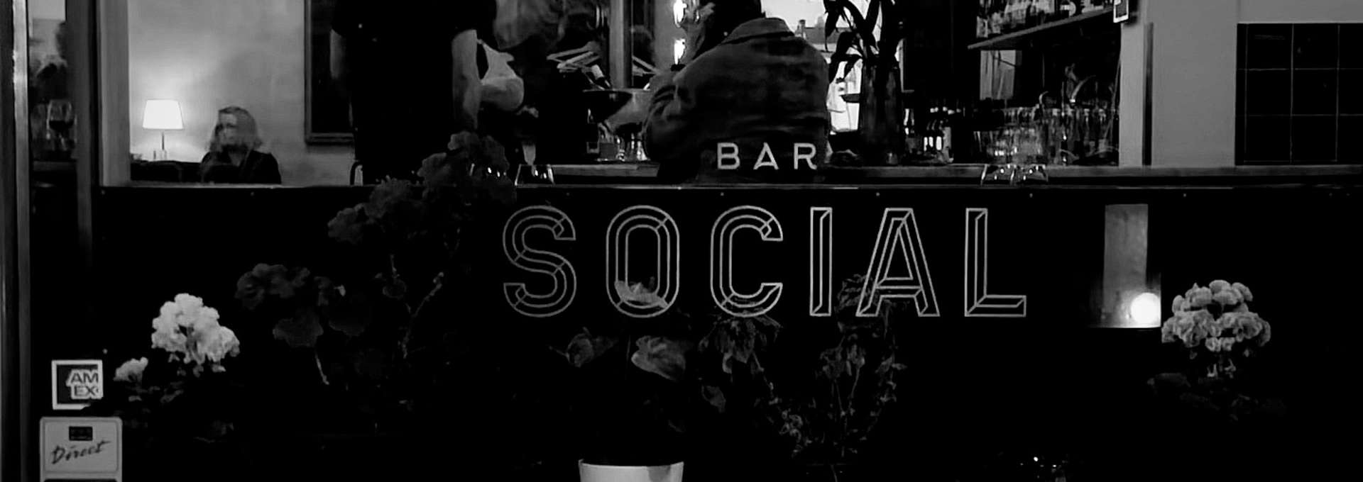 Svartvit bild tagen inne i en bar. Texten Bar social är tryckt på en spegel.