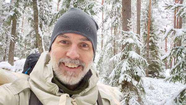 En leende man i svart mössta som står i en snötäckt skog.