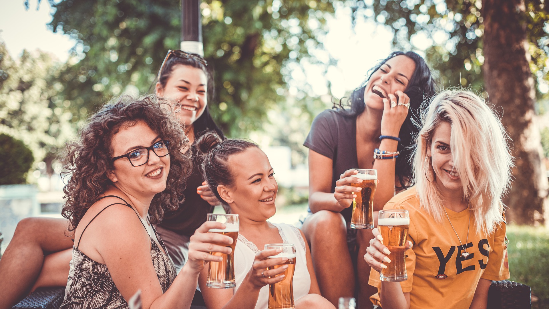Fem glada tjejer sitter på en uteservering en sommardag och dricker öl. 