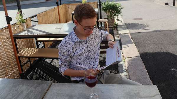 En ung man i ljus skjorta som sitter på en uteservering och läser ett magasin. Bredvid står ett glas rött vin.