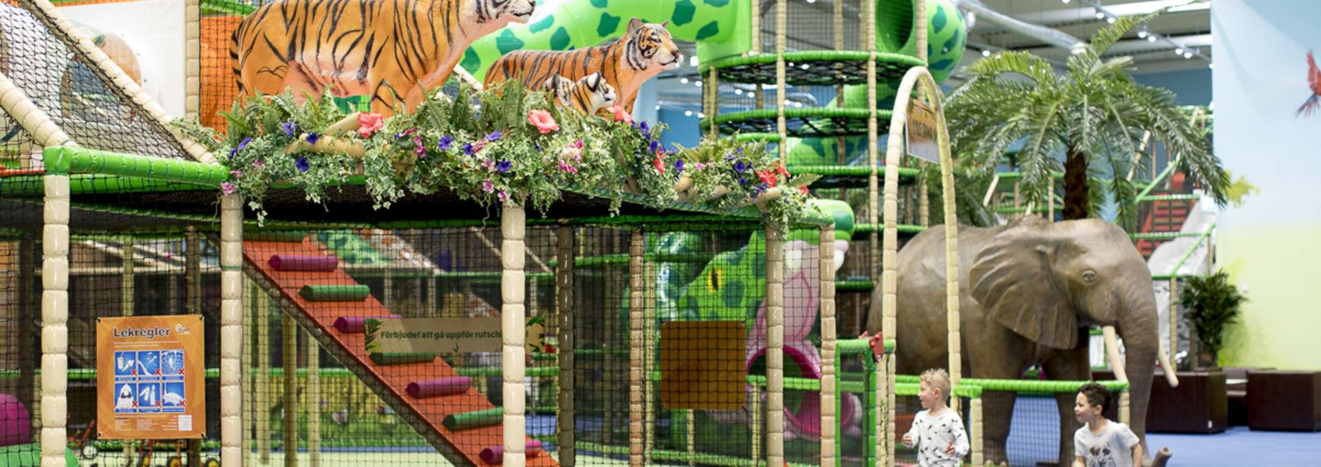 Två små pojkar springer och leker på Leos Lekland. På bilden syns stora färgglada klätterställningar och två tigrar och en stor elefant i plast.