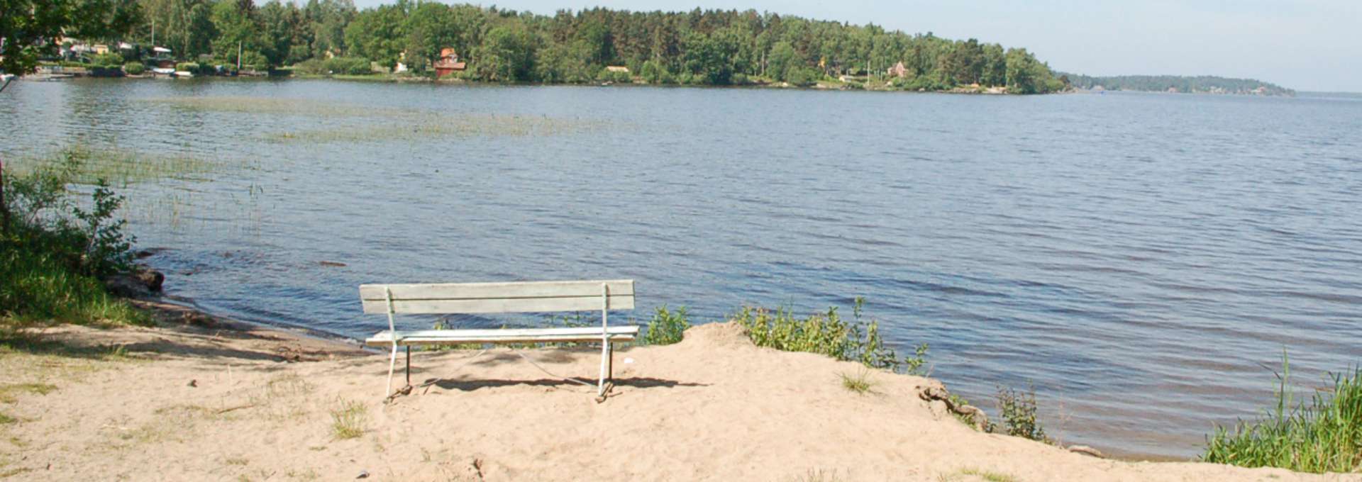 Badplats vid Mälaren med bänk, sandstrand och klarblått vatten