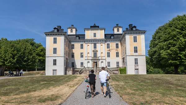 Mälsåkers slott i bakgrunden och två personer gåendes med cyklar syns på väg till slottet.