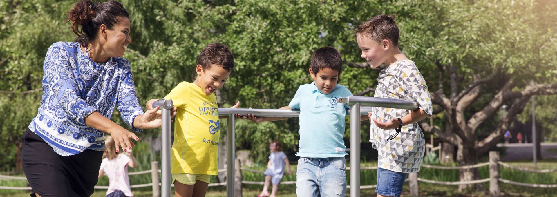 Barn leker på en lekplats en sommardag. De står på en rund ställning som går att snurra. en mamma hjälper till att ta fart. 