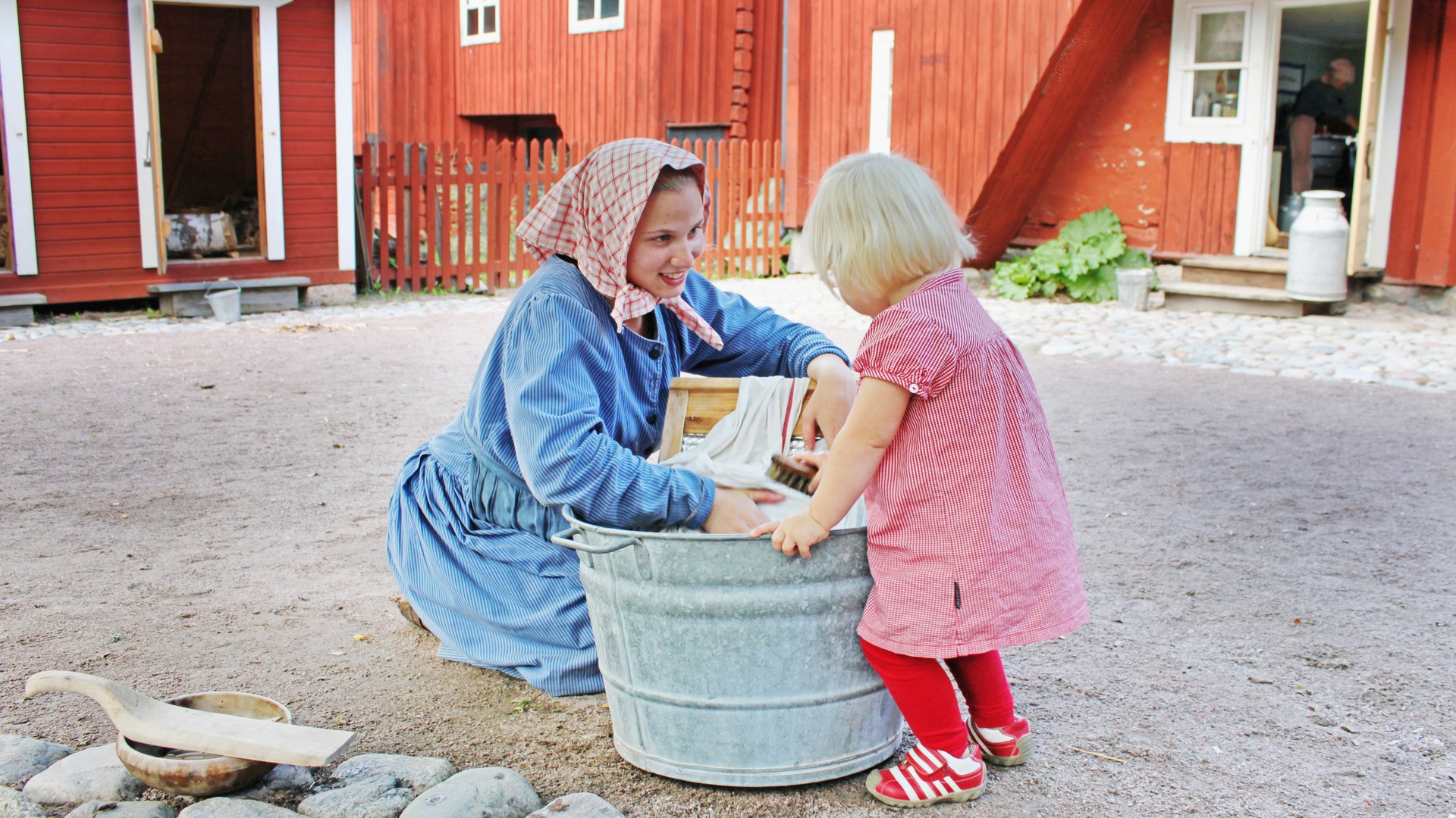 Gammaldags klädd kvinna tvättar kläder i en plåtbalja, en liten blond flicka tittar på.