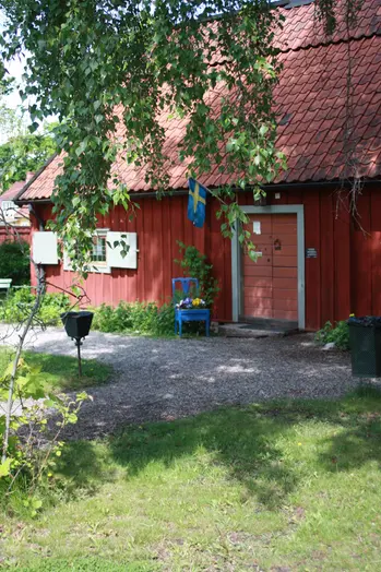 En gammalt rött hus med en svensk flagga bredvid dörren syns genom ett grönskande bladverk. 